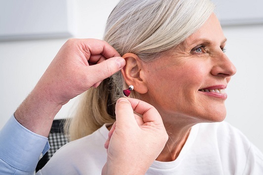 Ältere Frau bekommt ein modernes Hörgerät ins Ohr eingesetzt und lächelt zufrieden.